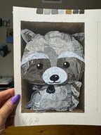 A hand with purple nail polish holds a square canvas painting of a raccoon made from crumpled silver foil, set against a white textured paper background. The painting is shown in an indoor setting with a wooden table surface below.
