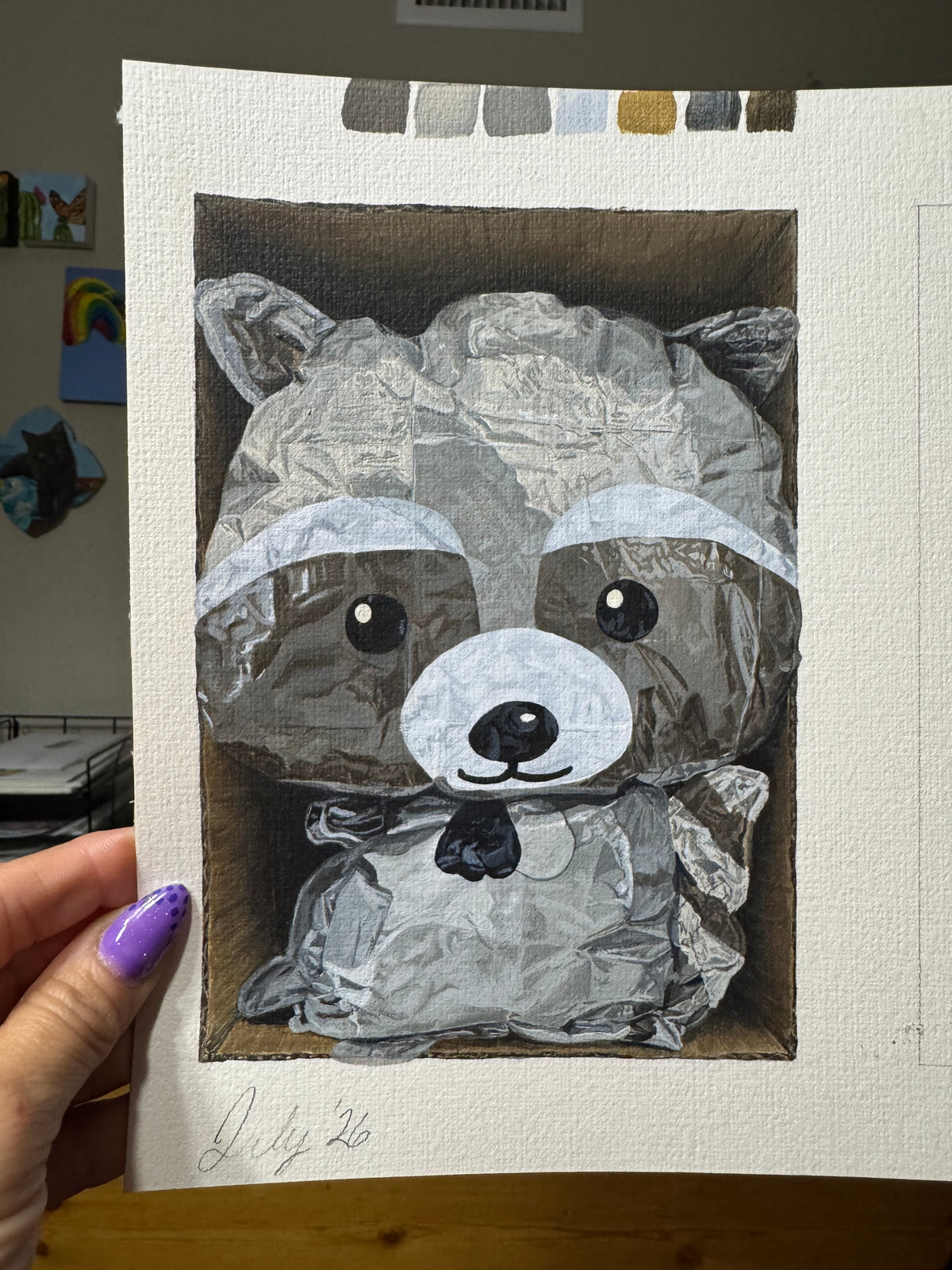 A hand with purple nail polish holds a square canvas painting of a raccoon made from crumpled silver foil, set against a white textured paper background. The painting is shown in an indoor setting with a wooden table surface below.